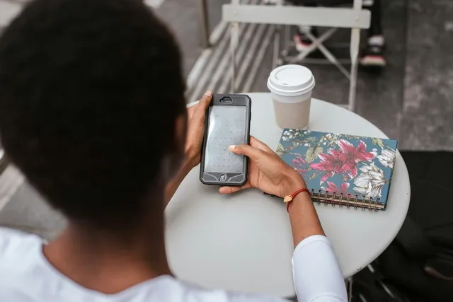 Person using a smartphone while sitting at a small table with coffee and a notebook
